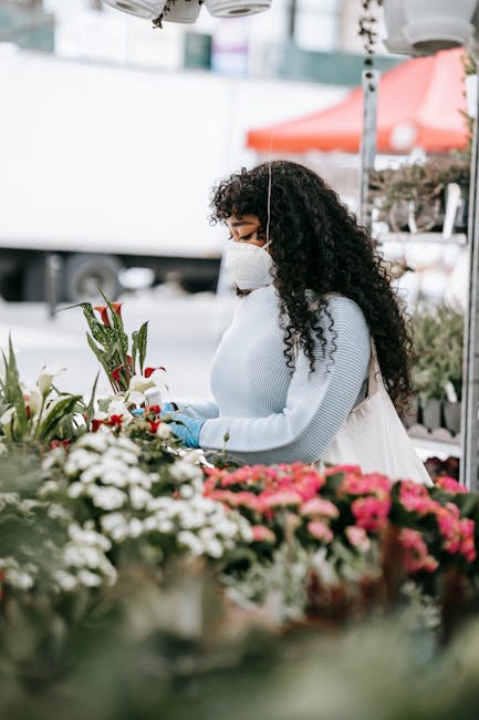 A woman with long, curly dark hair wearing a white face mask, light blue sweater, and blue gloves is arranging a bouquet of fresh flowers at an outdoor flower market. The bouquet features white lilies