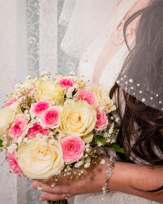 A bride's hand is shown holding a bouquet arrangement featuring large, creamy white roses and smaller pink roses with delicate white baby's breath accents. The bouquet is wrapped in light, translucent
