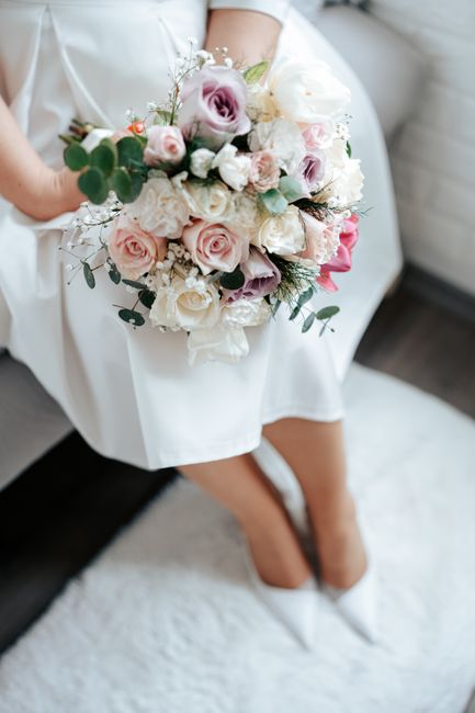 A bride dressed in a white wedding gown, sitting on a white textured rug, holding a large bouquet of fresh flowers. The bouquet features pale pink roses, white lisianthus, light purple eustoma, and sp