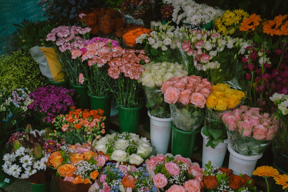 A vibrant display of fresh flowers arranged in various bouquets and pots, featuring roses in soft pink, white, and yellow, along with clusters of carnations in salmon and coral hues. Purple and white 