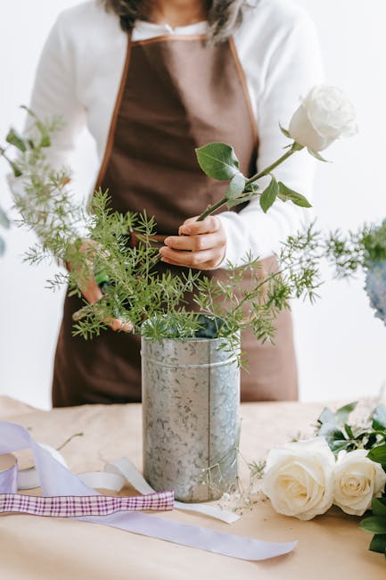 A person dressed in a white shirt and brown apron arranging a floral display, holding a single white rose with green leaves. The arrangement includes various green foliage placed in a silver metallic 