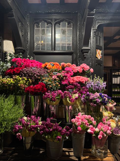 An arrangement of vibrant fresh flowers including pink, purple, orange, and red blossoms, displayed in tall, metallic vases on a dark wooden shelf in a floral shop setting. The flowers feature roses, 