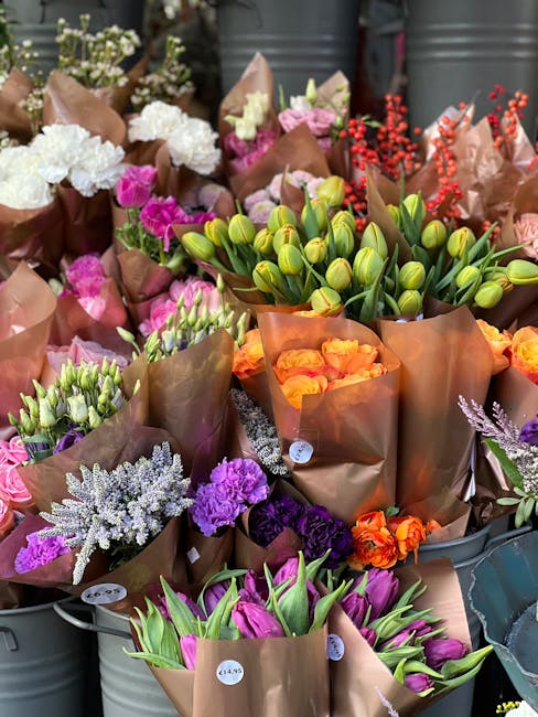 A vibrant display of freshly cut flowers arranged in brown kraft paper wrapping, including tulips in shades of purple and pink, orange roses, closed green tulip buds, and clusters of white, purple, an