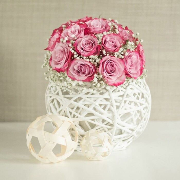 This image presents a tranquil tabletop vignette centred around a lush, dome-shaped bouquet of pink roses and delicate white baby's breath, the kind of arrangement you might see gracing a sunlit sitting room in Farnborough's older streets near North Camp. The bouquet is compact and rounded, with around fifteen soft pink roses nestled closely together so that their layered petals form a near-perfect sphere of colour. Each rose displays a graceful colour transition, beginning with a pale blush at the heart and gradually deepening to stronger pink and near-fuchsia hues along the outer edges, giving the impression of gentle warmth spreading outward. Interspersed through the roses are fine, branching stems of gypsophila, their myriad tiny white blooms creating pockets of softness and air between the larger flowers. This contrast of size and texture adds an almost cloud-like lightness around the more substantial rose heads, balancing romance with simplicity. The arrangement is anchored in a distinctive spherical vase crafted from white, woven strands that recall wicker or rattan, painted and shaped into an open lattice. The criss-cross pattern allows glimpses of shadow within the sphere, creating visual depth and giving the vase an almost sculptural quality. It stands on a smooth, immaculate white surface that gently reflects the light, adding a sense of freshness. To the left, two woven decorative spheres rest casually yet purposefully: both are made from thin natural-fibre strips in a warm beige tone, with the larger ball placed slightly behind the smaller one, echoing the woven motif of the vase. A softly textured wall in warm beige or cream provides a calm, neutral backdrop, similar to the soothing interiors of modern apartments near Farnborough Main station. The lighting is soft and even, with no harsh shadows, lending the scene a quiet, contemporary elegance that would suit a thoughtful gift, a Mother's Day surprise, or a refined touch of décor for a home or boutique office in the area.
