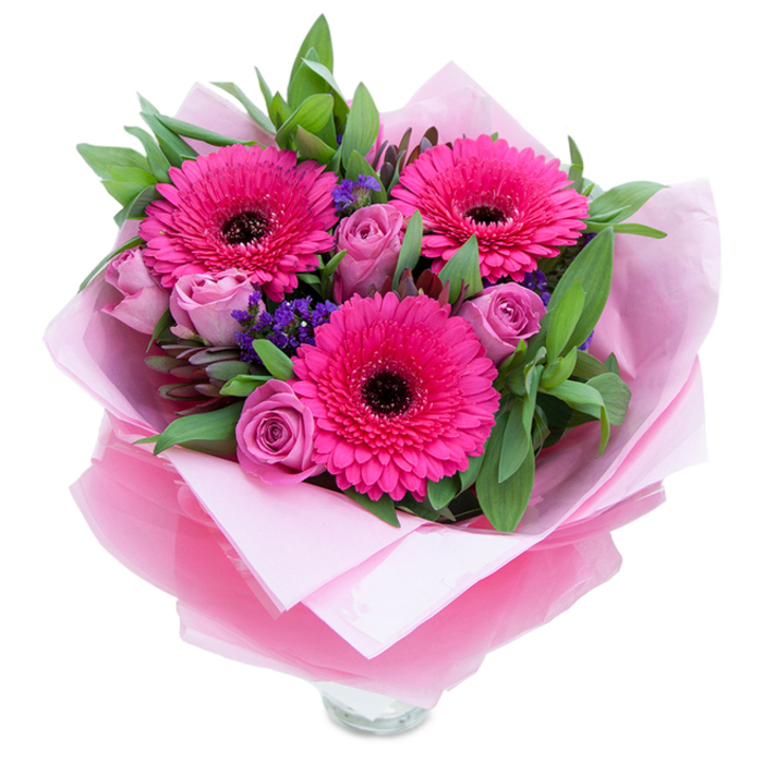 I see this bouquet as a graceful balance of boldness and softness: a trio of fuchsia Gerbera daisies unfurls with assertive colour and dark centres that anchor the eye, while surrounding lavender-pink roses introduce a quieter, more intimate rhythm with their tight, velvet-like petals. Scattered among the larger blooms are tiny clusters of purple statice (limonium), their papery texture and cool tones adding depth and a meadow-like looseness to the formal round shape. The greenery is abundant and purposeful - broad leaves that read as salal or eucalyptus give structure and a dewy, garden-sourced freshness that enhances the floral hues. The wrapping is part of the composition: an inner sheet of soft pale-pink tissue supplies a gentle tonal base, and over it a translucent fuchsia-pink cellophane, faintly crinkled, catches the light to add a whisper of glamour; expertly folded, it suggests a gift carefully prepared rather than a rushed impulse. The photograph reveals the top of a clear glass vase beneath the paper, a practical cue that the bouquet is meant to be enjoyed immediately on a mantel or bedside table, perhaps in a home not far from Farnborough town centre or the airport. Light and scent are implied - a light, sweet rose perfume offset by a fresh green hint from the foliage - making this an ideal choice for celebrations, Mother's Day, or a thoughtful 'just because' visit.