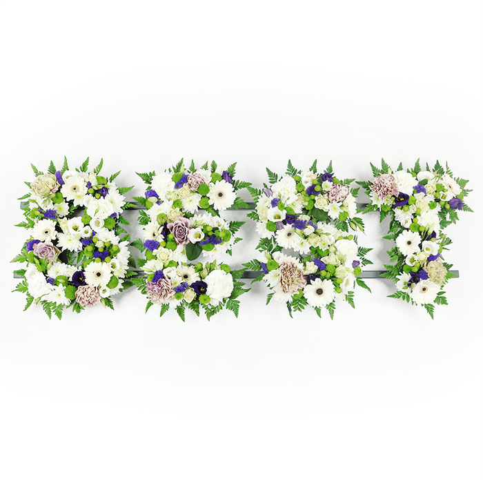 A carefully composed, horizontal memorial arrangement spells out BEST in bold capital letters, photographed on a pristine white backdrop to showcase every floral detail. Each letter is a concentrated mosaic of pristine white gerbera daisies with their dark central disks, soft white lisianthus and abundant white carnations that together create a serene, layered surface. Accents of soft mauve and lavender roses, along with deeper purple filler blooms, thread through the whites and lend a subtle, dignified warmth that deepens the emotional tone. Fresh green button chrysanthemums appear throughout, their compact shapes introducing an energetic freshness, while feathery green fern fronds trace and envelop each letter, producing a lush natural border and softening the graphic impact. The blooms are secured to a discreet dark grey or black support structure that peeks through in places, ensuring the letters remain clear and transportable for viewing at a service or memorial. The arrangement absorbs studio light in a way that highlights petal textures - the silky velvet of rose petals, the ruffled density of carnations and the taut, smooth faces of gerberas - and the overall scent profile is calm and floral, reassuring rather than overwhelming. I imagine this tribute being prepared in our Farnborough workshop near Princes Mead and set for delivery to a church or crematorium, chosen by loved ones who wish to express best-loved memories, deep respect and enduring remembrance. Crafted with care and local knowledge of Farnborough's funeral venues, it is both a visual statement and a tender, living message of honour.