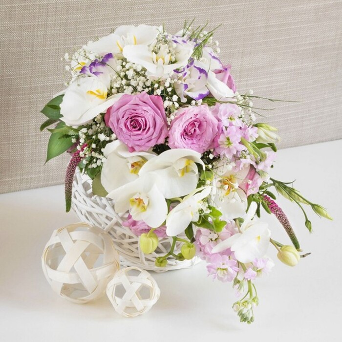 A beautifully composed floral arrangement in a round white wicker basket is photographed in detail, evoking the feeling of a refined wedding table set in a Farnborough venue. The basket brims with a carefully balanced mix of pink, white, and gentle lavender tones, dominated by lush pastel pink roses that are fully open, their layered petals forming perfect spirals that draw the eye to the centre of each bloom. Nestled alongside them, elegant white Phalaenopsis orchids unfold like butterflies, their broad, translucent petals framing small yellow-orange centres that introduce a hint of warmth and sophistication. Between these focal flowers, white Lisianthus blossoms add a soft, ruffled texture; some petals carry a delicate wash of purple along the edges, echoing the subtle lilac colours often seen in spring borders around Farnborough's residential gardens. Pale pink stock flowers rise and then gently trail from the arrangement, giving vertical interest and a graceful cascading line that would look beautiful down the centre of a wedding table. Fine sprays of white baby's breath are interspersed throughout, their tiny blossoms forming a misty, romantic veil around the larger blooms. Sprigs of fresh green foliage weave through the composition, with a few slender leaves and buds extending beyond the basket rim, suggesting natural growth and freshness, as if just arranged by an experienced local florist. The white wicker basket rests on a smooth white surface, accompanied by two decorative woven spheres in natural wood tones that echo the basket's texture and add a contemporary, organic detail. Behind, a softly textured beige fabric backdrop keeps the scene calm and neutral, allowing the colours and intricate petal details to stand out. The overall effect is one of gentle luxury, tenderness, and celebration, perfect for wedding receptions, anniversary dinners, or elegant gatherings across Farnborough and nearby areas.