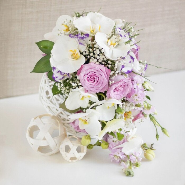 A sophisticated floral arrangement is carefully styled as a wedding-ready centerpiece, positioned on a crisp white surface that reflects the soft, even lighting. In the centre of the composition, immaculate white Phalaenopsis orchids command attention, each elegant bloom with broad, satiny petals and intricate yellow-gold markings in the throat that lend an air of luxury and poise. Surrounding these orchids, layers of pastel pink, lilac, and lavender roses create a plush, romantic collar; some roses appear tightly cupped, while others are fully open, displaying delicate, velvety spirals of petals that suggest both delicacy and abundance. Among them, daintier blossoms-likely lisianthus or eustoma-introduce subtle variation, their white or softly tinted petals edged with pale purple, contributing to a multi-dimensional, almost painterly texture. Fine sprays of white baby's breath filter between the larger flowers like a soft veil, filling gaps and adding an ethereal, clouded effect. A few stems of trailing, light purple blooms, perhaps stock or similar, flow gently downward, drawing the eye towards the base and giving the design a naturally cascading rhythm. All of this is gathered into a striking white spherical vase, its open, lattice-like weave suggestive of woven rattan or decorative twine, adding a touch of handcrafted sculpture that would look perfectly at home on a reception table at a Farnborough country house or near the quiet residential streets off Cove Road. Beside the vase are two decorative cream-toned spheres, woven in the same open pattern, one larger and one smaller, echoing the motif and adding visual balance. The background is a softly textured neutral beige, gently blurred to keep full attention on the fresh blooms and intricate florist artistry.