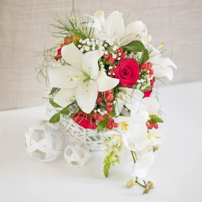 A luxurious floral display sits serenely on a smooth white surface, illuminated by soft, even light that emphasizes every petal and weave. The arrangement is cradled in a round, open-weave white container, its intricate basket pattern giving a handcrafted, contemporary feel often favoured by couples planning weddings around Farnborough's elegant hotels and country venues. Rising from this delicate framework, pristine white oriental lilies command attention, each bloom wide open, with thick, velvety petals and golden-tipped stamens that hint at a light, powdery fragrance. Tucked among the lilies are opulent red roses, their rich, velvety petals layered tightly, creating deep, romantic shadows of crimson that speak of passion and celebration. Between these focal blooms, fine sprays of white gypsophila, or baby's breath, appear like soft drifting clouds, providing a gentle, airy contrast to the larger flowers. Fresh green foliage and wispy, feathery greens add dimension and a sense of lush movement, reminiscent of a well-tended Farnborough garden after rain. Scattered within the bouquet are glossy red hypericum berries, their rounded forms bringing a hint of seasonal festivity, perfect for a Christmas wedding breakfast or winter anniversary dinner. From the right side of the basket, a cascading spray of white phalaenopsis orchids pours gracefully downward, their arching stems carrying multiple blooms and a few unopened buds, each flower with a soft yellow and pink heart that adds subtle warmth. To the left on the tabletop, two or three white decorative spheres made from matching woven material echo the container's texture, giving the entire scene a cohesive, curated look. The result is a refined, romantic centrepiece that would suit an intimate wedding reception near Farnborough town centre or an elegant dinner party at home, exuding calm luxury and understated joy.