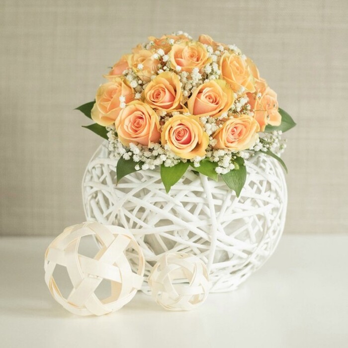 A refined floral composition showcases a full, domed cluster of peach and apricot roses, tightly packed so that nearly every inch of the bouquet is filled with layered petals. Each rose shows a soft gradient of colour, from richer golden apricot at the centre to a delicate, almost blush-peach at the edges, evoking the warmth of a late afternoon sky over Farnborough's parks. Threaded through the roses are generous sprigs of white baby's breath, their tiny blossoms forming a fine, cloud-like veil that wraps the bouquet in softness. Beneath the roses, glimpses of rich, dark green foliage peek through, giving structure and a natural foundation without overtaking the gentle palette. The arrangement sits in a distinctive, spherical white vase created from slim wicker or rattan strands, woven into an open, crisscrossing lattice. This airy structure allows light to pass through, creating intricate shadows on the pristine white tabletop below. In the foreground, two decorative spheres made from natural-toned, woven strips-one larger, one smaller-echo the same texture and bring a subtle, sculptural element to the scene. The background is a muted light beige or off-white wall with subtle vertical texturing, ensuring there are no distractions from the flowers while still offering visual interest. Soft, even daylight illuminates the entire setting, catching on the wicker and highlighting the velvety surface of each petal. The result feels serene and sophisticated, like a carefully styled corner of a modern Farnborough home near Southwood or a boutique hotel lobby. It is easy to imagine this arrangement as a centerpiece for a wedding breakfast, an engagement dinner, or a beautifully staged property, with a local florist ready to deliver a similar design across the GU14 area.