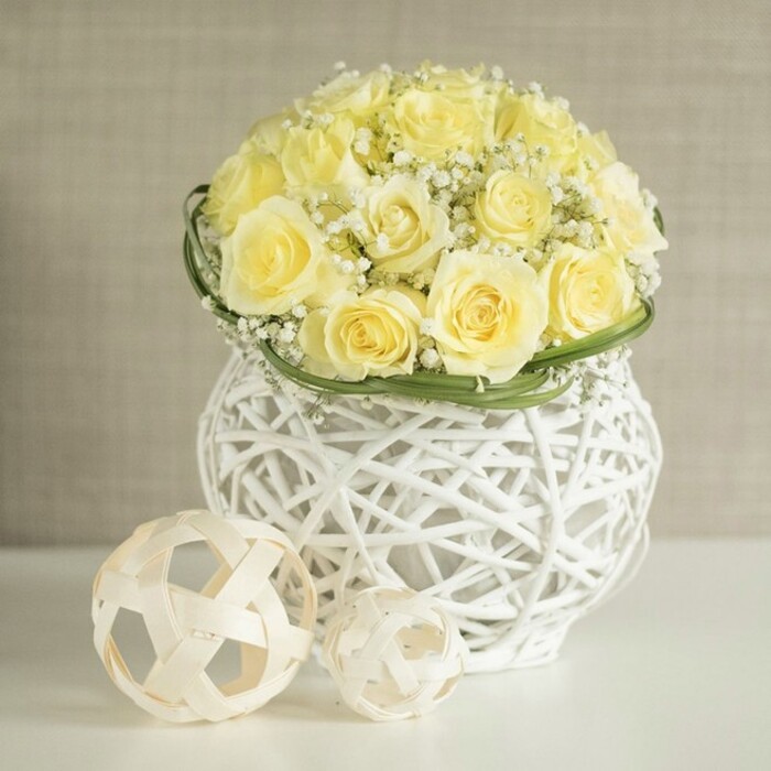 A refined floral display, ideal for a wedding table in Farnborough, is captured against a softly blurred, warm beige background that keeps all attention on the artistry of the design. At the centre is a lush, rounded bouquet of pale yellow roses, each head full and perfectly opened so that the petals spiral outward in gentle layers. The roses sit so close together that they form a smooth, cushion-like canopy of colour, their creamy yellow tones suggesting early morning sunlight. Between their rounded forms, delicate sprigs of white baby's breath emerge, thousands of minuscule blooms creating a gauzy veil that lightens the entire arrangement. Just below this floral dome, a precise band of slender green foliage circles the bouquet, almost like a ribbon of living leaves, marking a graceful transition from flowers to container. The vase itself is a striking white piece, built from intricately woven strands that mimic rattan or wicker, looping and intersecting to create a basket-like lattice with generous openings. This open-work structure allows light to pass freely, casting subtle shadows and giving the vessel a sculptural, modern feel, as though it belongs in a stylish Farnborough wedding venue or a chic home near Cove or Southwood. Resting casually on the shining white tabletop are two natural woven balls in complementary light wood tones, one slightly larger and positioned forward, the other tucked beside it. Their open, geometric weave mirrors that of the vase, tying all the elements together. The arrangement feels joyful yet soothing, perfect for decorating reception tables, intimate anniversary dinners, or spring celebrations, and reflects the kind of careful craftsmanship a local Farnborough florist would prepare for same-day delivery and setup.