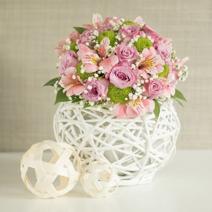 Capturing the elegant spirit of Farnborough's welcoming homes, this stunning floral arrangement creates a visual and emotional focal point for any gathering. Against a soft, light beige backdrop that speaks to tranquil Farnborough mornings, the arrangement is lavishly composed of lavish pastel pink and lavender roses, their lush, velvety petals radiating delicate charm. They mingle elegantly with trumpet-shaped pink alstroemeria marked by subtle streaks of yellow and deep pink, their blooms nestling between flashes of vibrant lime green button chrysanthemums. Wisps of baby's breath, pure white and airy, fill the negative space, lending an almost weightless quality to the ensemble. The floral display is expertly gathered in an impactful white vase, notable for its intricately woven, open-lattice design-fashioned from slender strips in a manner that captures both rustic artistry and modern flair. Its rounded, sturdy base provides the perfect foundation for the fountain of blooms above. On the pristine tabletop to the left lie two woven decorative spheres, each subtly echoing the craftsmanship of the vase but varying in size for natural asymmetry and interest. These pale, rattan-like orbs suggest both creativity and comfort, calling to mind bespoke touches found in Farnborough's boutique shops. The scene as a whole evokes a sense of sophisticated celebration-ideal for a wedding table at a local venue off Lynchford Road, or as an exquisite surprise arranged for delivery by Flower Delivery Farnborough. This centerpiece speaks of freshness, artistry, and local pride, a testament to the thoughtful beauty that defines community celebrations here.