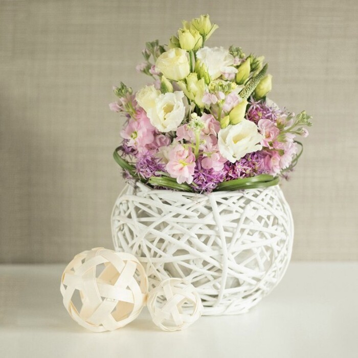 This elegantly curated floral centrepiece captures the serene charm of Farnborough springtime in every detail. At the core, creamy white and pastel yellow eustoma unfurl their sumptuous ruffled petals, exuding a gentle luminosity. Interspersed are spires of soft pink stock, their slender shapes rising with poise-much like the blossoming trees found along the paths of Moor Road. Adding dimension beneath, miniature clusters of vivid purple allium or aster create a textured foundation, their depth enhanced by the fresh vibrancy of deep green foliage encircling the base, all thoughtfully arranged by skilled hands familiar with the tastes of Farnborough residents. The florals are presented in a unique spherical vase, expertly woven from painted white rattan in an open-work pattern, lending a striking yet lighthearted air. Adjacently placed, two decorative sphere accents constructed from pale, natural wood in intricate basket-weave designs orbit the vase. Their organic shapes mirror the artistry of the surrounding bouquet, creating harmony within the display. The ensemble rests against a warm, gently textured beige wall, subtly reminiscent of Farnborough's inviting interiors or boutique cafes. Light dances softly across the scene, evoking a sense of calm, freshness, and refined celebration-an atmosphere ideal for a wedding table at a local venue, a spring luncheon, or sophisticated home décor. This arrangement, by Flower Delivery Farnborough, beautifully weaves local charm, visual delight, and florist expertise into a centrepiece that embodies both elegance and heartfelt welcome.