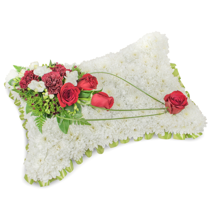 This poignant rectangular floral cushion is a study in contrast and calm: hundreds of small white chrysanthemum heads are densely arranged to create a soft, velvety plane that evokes purity and rest, while a narrow band of light green ribbon or foliage edges the form and offers a fresh, reassuring line. Nestled at the top left, a composed spray of deep red roses and rich burgundy carnations provides a solemn, warm counterpoint, their velveteen petals and ruffled textures speaking of love and lasting remembrance. Scattered among them are tiny white accent blooms-freesias or spray chrysanthemums-that add delicate lightness and a faint, clean perfume. Underpinning the floral cluster, abundant greenery, including slender ferns and fuller leaves, deepens the tonal palette and frames the blooms. From this left-hand cluster, graceful stems of bright green bear-grass or decorative foliage arc diagonally across the white chrysanthemum surface, drawing the eye and creating a sense of movement that leads to a single, perfectly centred red rose resting near the lower right corner. That solitary rose reads as a gentle offering, a personal touch within a classic bereavement design. I picture this arrangement being chosen for services held at local chapels or for placement at memorials in Farnborough's quieter neighbourhoods-delivered with care and designed to convey dignity, comfort, and heartfelt sympathy.