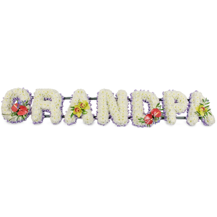 This image shows a dignified, horizontal floral tribute spelling GRANDPA in large, upright block letters, crafted with the precision and care you would expect from an experienced Farnborough funeral florist. Each individual letter is built up from a dense bed of small, snow-white chrysanthemums, their rounded petals forming a plush, almost velvety carpet of blooms that reads clearly from a distance. The flowers are arranged closely so no gaps in the greenery can be seen, creating a pure white canvas that suggests innocence, remembrance, and peace. Along the base of every letter, a soft border of pale lavender ribbon or trim adds a gentle line of colour, framing the design and giving the tribute a subtly finished, tailored look. Supporting bars in dark green run along the length of the piece, linking the letters into one continuous, stable display that would sit securely atop a coffin or at the front of a chapel in Farnborough or nearby villages. To break the expanse of white, rich accent sprays are placed with intention: on the G and D, clusters of deep red anthuriums draw the eye, their characteristic heart-shaped, waxy spathes and bold yellow spadices emerging from a nest of lush foliage, perhaps with small hints of complementary purple filler flowers. On the first A and the final A, elegant yellow-green orchids cascade gently over the chrysanthemums, their arching stems and intricate blooms bringing an air of refinement and lasting beauty. This bespoke tribute feels deeply personal, a floral headline honouring a grandfather's role at the centre of family life, ready to accompany a service at the local church or Farnborough crematorium with quiet strength and affection.
