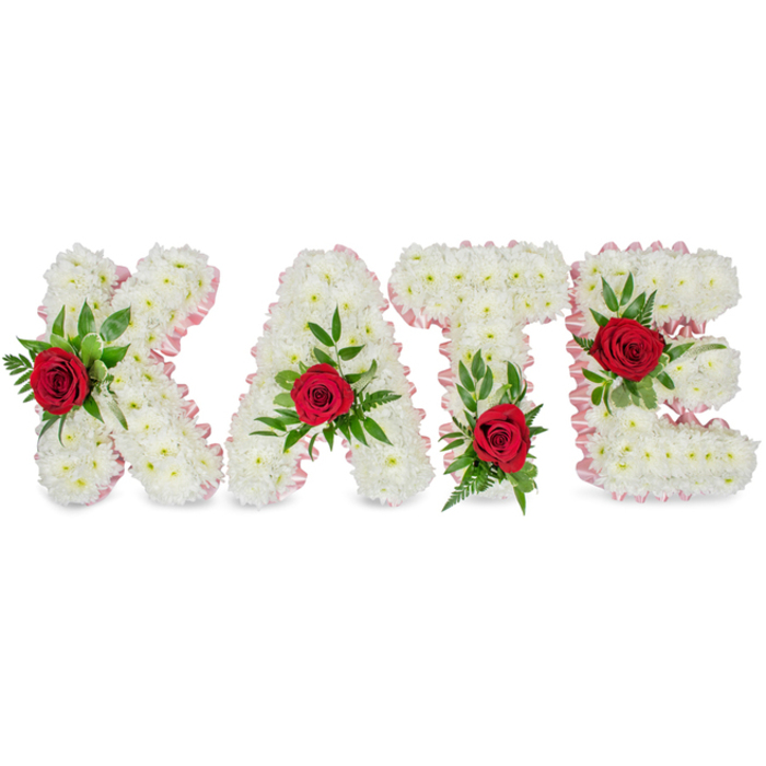 This image presents an elegant, personalised floral tribute forming the name KATE in pronounced block letters, each letter sculpted from a voluminous bed of small, pure-white chrysanthemum blooms. The chrysanthemums sit densely packed to create smooth, cloud-like contours that communicate calm and reverence, set cleanly against a white studio background so every texture and hue reads clearly. The edges of each letter are gently finished with a delicate pale pink satin ribbon that adds a refined border and a whisper of warmth to the composition. Each letter is further embellished with a single, vivid red rose: on the K the rose is arranged toward the lower section of the left upright and softened by surrounding greenery; on the A the rose is placed near the top peak and cradled by fern fronds; the T receives its red accent at the base of the left side of the horizontal bar; and the E bears its bloom along the centre horizontal arm, each rose framed by layers of lush, glossy leaves and fine, airy foliage. The overall effect balances purity and passion - the white chrysanthemums symbolising remembrance, the red roses declaring love - while the green elements introduce life and resilience. Imagine this piece in soft morning light, its textures and gentle scents appropriate for a funeral, memorial service or private commemoration in Farnborough or a nearby chapel.