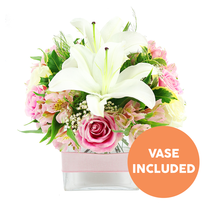 An elegant floral display featuring white Oriental lilies and shades of pink roses is carefully styled in a low, square glass vase and photographed against a bright white background, giving it the polished appearance of a gift just delivered somewhere in central Farnborough. Two fully open white lilies dominate the centre, their broad petals arching gracefully and revealing prominent reddish-brown stamens that draw the eye. Around them, clusters of pink roses form a rounded, almost dome-like silhouette; some roses retain their tight, spiralled centres, while others open wider to showcase layers of satin-soft petals that suggest warmth, gratitude, and gentle romance. Interlaced among the roses are smaller blooms of alstroemeria in pale pinks and soft peach, each floret marked by subtle speckling and fine veins of yellow that lend a painterly, detailed texture. Fine clouds of white gypsophila nestle in between, softening any gaps and giving a light, airy feel, as if the bouquet is breathing. A mix of deep green foliage provides a lush contrast, including broad, polished leaves and more delicate, feathery greens reminiscent of the varied planting you might see in local Farnborough gardens or along the paths by the Basingstoke Canal. The clear vase reveals straight, fresh stems, and around its centre sits a refined pale pink ribbon, tied smoothly to echo the rosy tones above without overpowering them. In the bottom right corner of the image, a round, orange graphic with bold white capitals spells out VASE INCLUDED, clearly indicating that the arrangement is complete and ready to place on a table or desk. The overall mood is one of understated elegance and freshness, suitable for birthdays, thank-you gestures, or simply brightening a Farnborough home. Crafted with a florist's eye for balance and texture, it feels both modern and timeless.