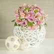 Bathed in gentle, natural light and nestled on a flawless white surface, this handcrafted arrangement is a celebration of both florist artistry and the quiet elegance of Farnborough. The voluminous bouquet unfolds in a spectrum of pastel pink and lavender, anchored by classic velvety roses, their petals soft and abundant. Sprigs of pink alstroemeria, marked with distinctive streaks and subtle yellow touches, add an air of refined delicacy, while lively lime green button chrysanthemums punctuate the arrangement with their bright, compact forms. Airy sprigs of baby's breath are artfully scattered throughout, lending a dreamy lightness to the display, while scattered deep green leaves create a sense of grounded freshness. Cradling it all is a white vase with an intricate, lattice-like weave, each slender strip crossing and curving with intentional care-a piece that would not feel out of place in the cozy interiors of Farnborough's cafes along Queensmead. This contemporary yet rustic vase rests securely at the center, while to its left, two natural-toned decorative spheres-crafted from light woven material in varying openwork patterns-echo the vase's design and enrich the table's visual rhythm. Their placement and shape create balance and movement, a subtle nod to the harmonious flow found in local community gatherings. This scene is perfect for inspiring wedding décor or marking special moments in Farnborough, where thoughtful design and fresh, vibrant blooms transform everyday occasions into cherished memories.