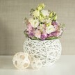 Bathed in the soft, diffused daylight often found in Farnborough's quieter neighbourhoods, a handcrafted floral display takes centre stage atop a gleaming white tabletop. Central to the composition is a rounded vase, masterfully constructed from strands of painted white rattan, its open latticework blending artisanal skill with contemporary design-reflecting the modern yet grounded style popular throughout the Farnborough area. Overflowing from this vessel, creamy and pale yellow eustoma unfurl with lush, ruffled petals, while delicate pink and lavender matthiola threads vertically between, recalling the gentle blooms spotted in local spring gardens. Framing the structure, vivacious purple filler flowers-likely allium or aster-provide a burst of colour, balanced by a neat ruffle of rich, green foliage encircling the arrangement. Completing the visual story, two decorative accents fashioned from light-hued wood and woven into sphere shapes rest gracefully at the side, their patterns echoing the openness of the vase and infusing a relaxed, welcoming tone. The scene is set against a subtle, beige backdrop, its soft texture reminiscent of Farnborough's charming home interiors or boutique venues. The air seems filled with the scent of fresh petals, while the overall impression evokes tranquility, elegance, and the quiet excitement of a special occasion shared with friends and family. This centrepiece would feel equally at home gracing a spring wedding table in Farnborough or offering a touch of handcrafted warmth to a local home, embodying both artisanal creativity and the spirit of community celebration.